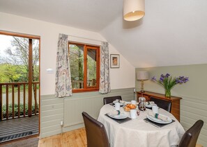 A dining area with a table set for two, a window with curtains, a lamp, and a vase with flowers.