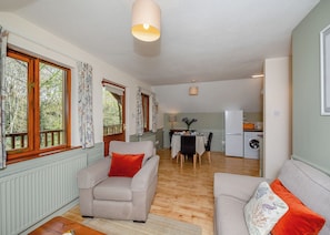 Una sala de estar acogedora con sofá, una mesa de comedor con sillas, una zona de cocina con refrigerador y lavadora, y ventanas amplias con cortinas.