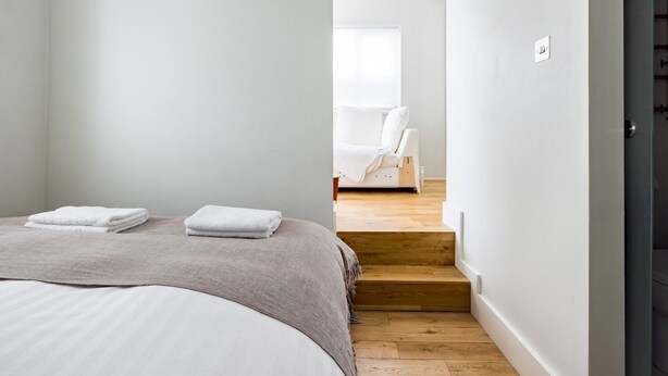 A bedroom with a bed, a bedside table, and a chair in the adjacent room. - Chic Apartment, Steps away from King's Rd Chelsea - Pass the Keys (London)