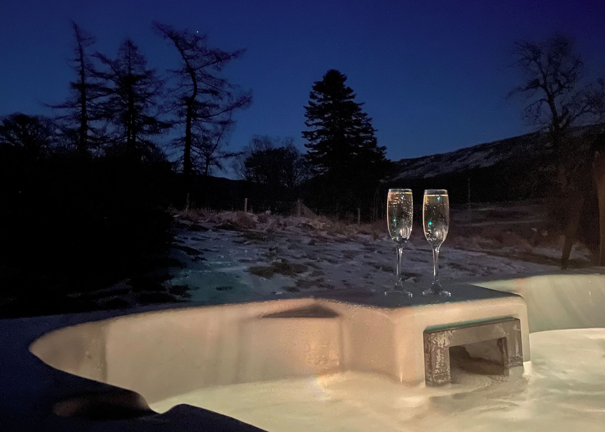Un bain à remous avec deux coupes de champagne, entouré d’un paysage enneigé et d’arbres, au crépuscule.