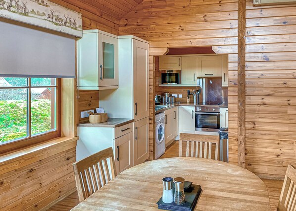 Interior de una cabaña de madera con una mesa de comedor, zona de cocina y vistas al exterior.