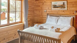 Une pièce aménagée comme un chalet en bois, avec un lit, une fenêtre avec des rideaux, un tableau encadré au mur et une petite table.