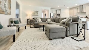 A modern living room with a sectional sofa, ottoman, and coffee table. There is a ceiling fan, a map on the wall, and a kitchen in the background. - Lots of amenities. Open Floor plan. Perfect for families and groups!  (Auburn)