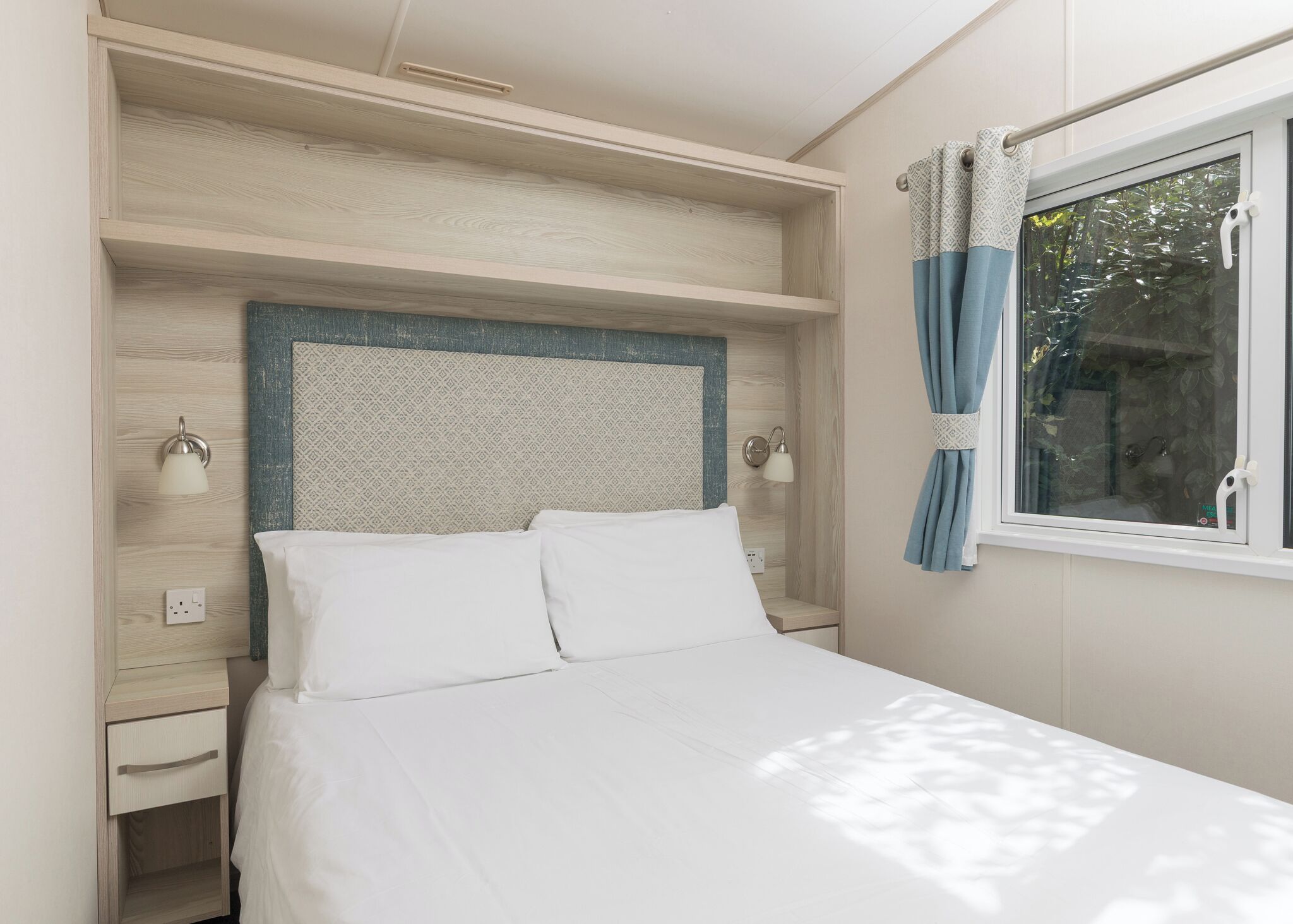 A bedroom with a bed, bedside lamps, a window with curtains, and a built-in shelf above the bed.