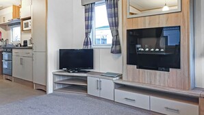 Un espace de vie compact comprenant un coin cuisine, un téléviseur à écran plat fixé à un mur en bois et une fenêtre avec des rideaux.