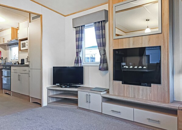 A compact living space with a kitchenette, a flat-screen TV mounted on a wooden wall, and a window with curtains. - Tollerton Caravan WF (Tollerton, York)