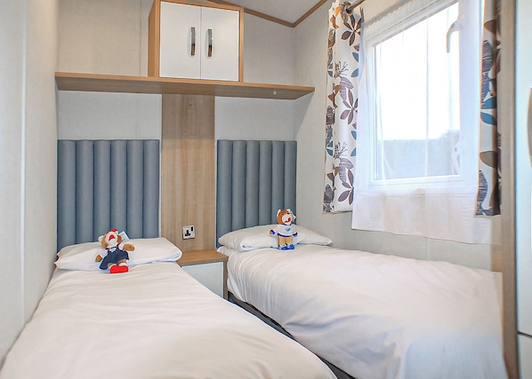 A bedroom with two beds, a wardrobe, a window with curtains, and a teddy bear on one bed.