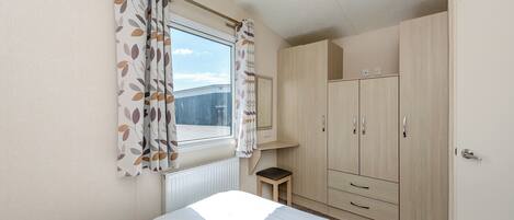 A bedroom with a bed, a window with curtains, a radiator, a small stool, and a wardrobe.