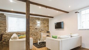 Un salon avec un mur en pierre, un plafond avec une poutre en bois, un téléviseur à écran plat et deux canapés blancs.