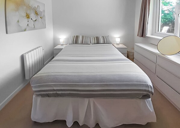 Une chambre à coucher comprenant un lit, des tables de chevet, une commode, un radiateur et une fenêtre avec des rideaux.