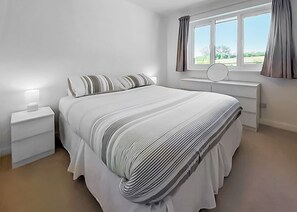 A bedroom with a bed, a bedside table, a nightstand, a window with curtains, and a view of greenery outside.