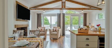 Een ruime kamer met een houten plafond, een eethoek met een tafel voor vier personen, een zithoek met twee stoelen, en een keukenblad met een spoelbak en opbergladen.