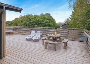 Eine Holzterrasse mit Liegestühlen und einem Picknicktisch, umgeben von einem Holzzaun und Grünpflanzen.
