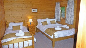 A wooden cabin bedroom with two beds, a nightstand, a lamp, and a framed picture on the wall. - Spacious timber lodge with one double and one single in one room. (Kentisbeare, Nr Cullompton)