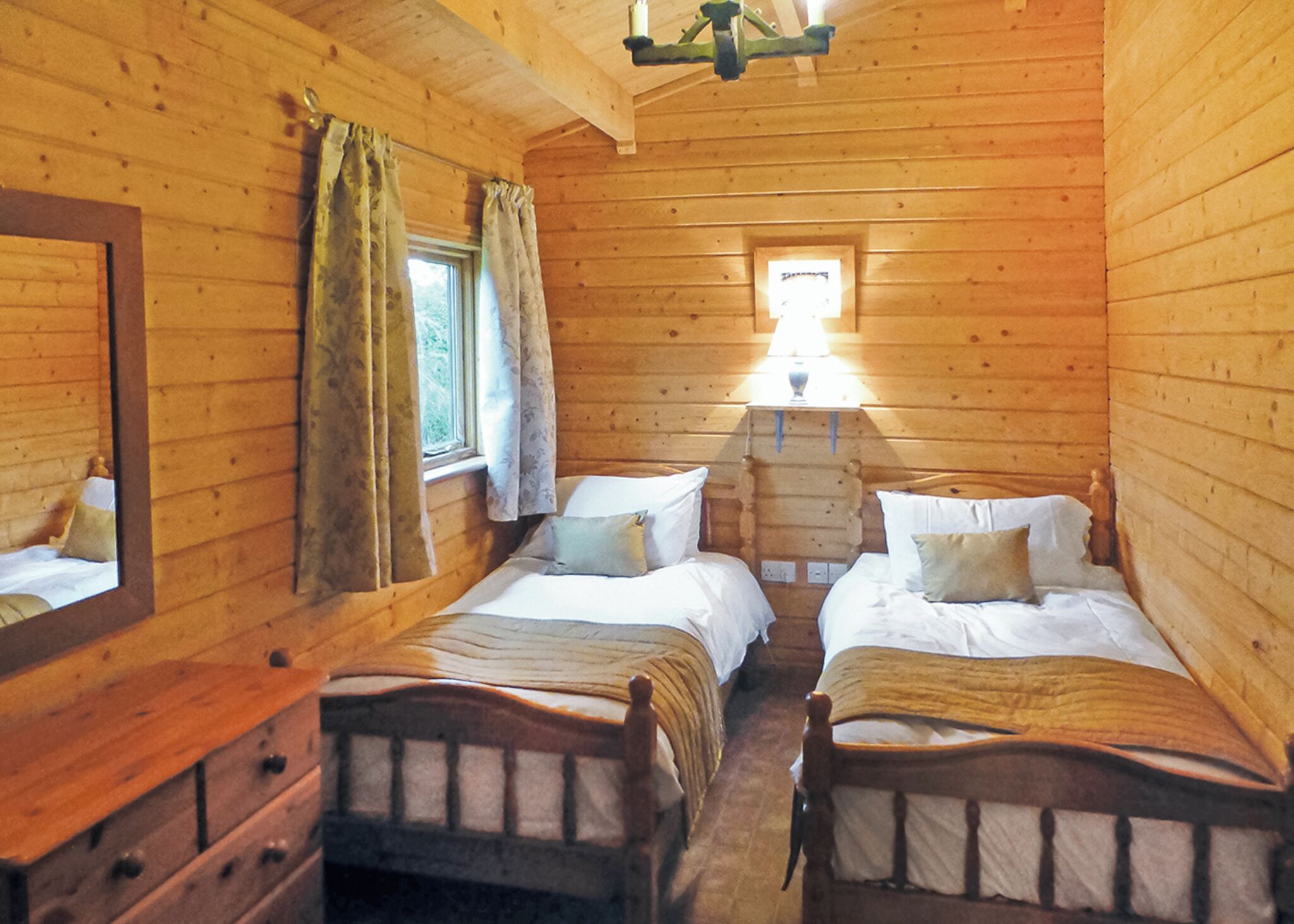 Une chambre de style cabane avec deux lits en bois, une commode, un miroir et une fenêtre avec des rideaux.