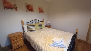A neatly made bed with a floral bedspread, a wooden nightstand with drawers, a bedside lamp, and three wall-mounted paintings. - Cuckoo Tree House, Glengarriff, Beara Peninsula (Glengarriff)