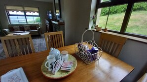 A dining table with a tea set, a basket of items, and a view of a grassy yard through large windows. - Cuckoo Tree House, Glengarriff, Beara Peninsula (Glengarriff)