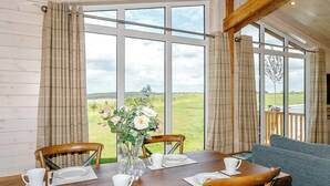 A dining area with a table set for two, a vase with flowers, and a view of a green landscape through large windows.