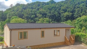 Ein kleines, eingeschossiges Gebäude mit geneigtem Dach, einer Holzterrasse und einem Garten mit Blumen.