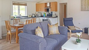 Ein Wohnzimmer mit Sofa, einem Esstisch, Stühlen, einer Küchenzeile mit Schränken und Geräten sowie einem kleinen weißen Tisch mit einer Pflanze.