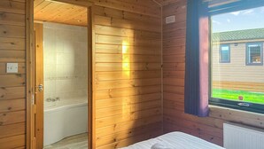 Une chambre dans un chalet en bois, avec un lit, une salle de bain dotée d’une baignoire et une fenêtre donnant sur une maison.