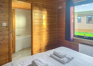 A wooden cabin room with a bed, a bathroom with a bathtub, and a window with a view of a house.