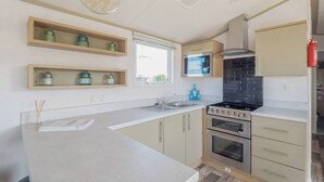 A modern kitchen with a central island, built-in microwave, oven, and gas stove. There are wooden shelves above the island and a window providing natural light. - Stylish and comfortable holiday home featuring your own private hot tub. (Laurenkirk)