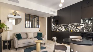 Una sala de estar moderna con sofá, mesa de centro y sillas, una cocina con armarios oscuros y salpicadero de mármol, y un espejo redondo en la pared.