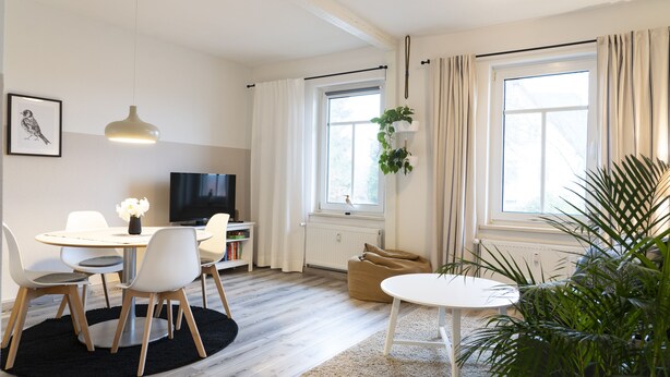 A modern living room with a dining area, a television, and a large window with curtains.