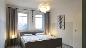 A bedroom with a bed, two bedside lamps, a window with curtains, a door, and a wall-mounted picture.