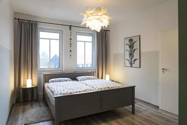 Een slaapkamer met een bed, twee bedlampen, een raam met gordijnen, een deur en een aan de muur hangend schilderij.