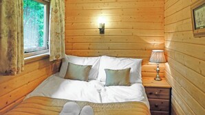 A wooden cabin bedroom with a bed, pillows, a nightstand, a lamp, and a window with curtains.