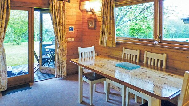 A wooden cabin interior with a dining table, chairs, curtains, and a view of the outdoors.