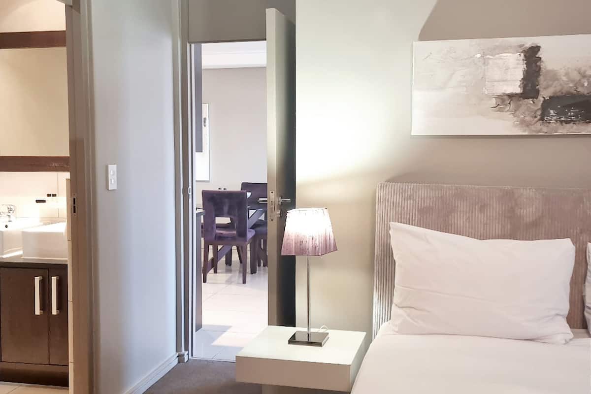 A hotel room with a bed, bedside table, lamp, and a view of the dining area.