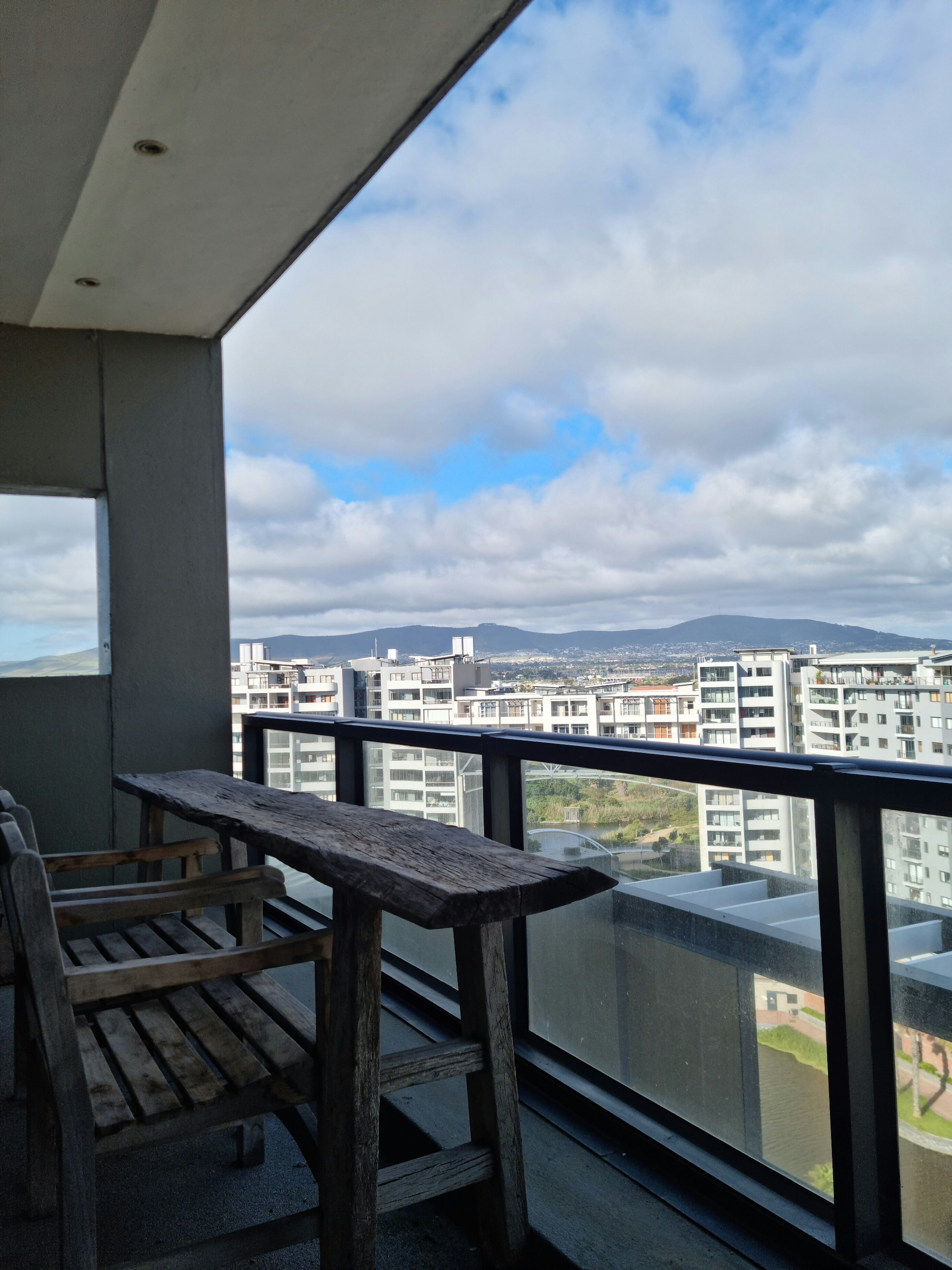 A balcony with a wooden bench and a view of a cityscape with buildings and mountains in the distance.