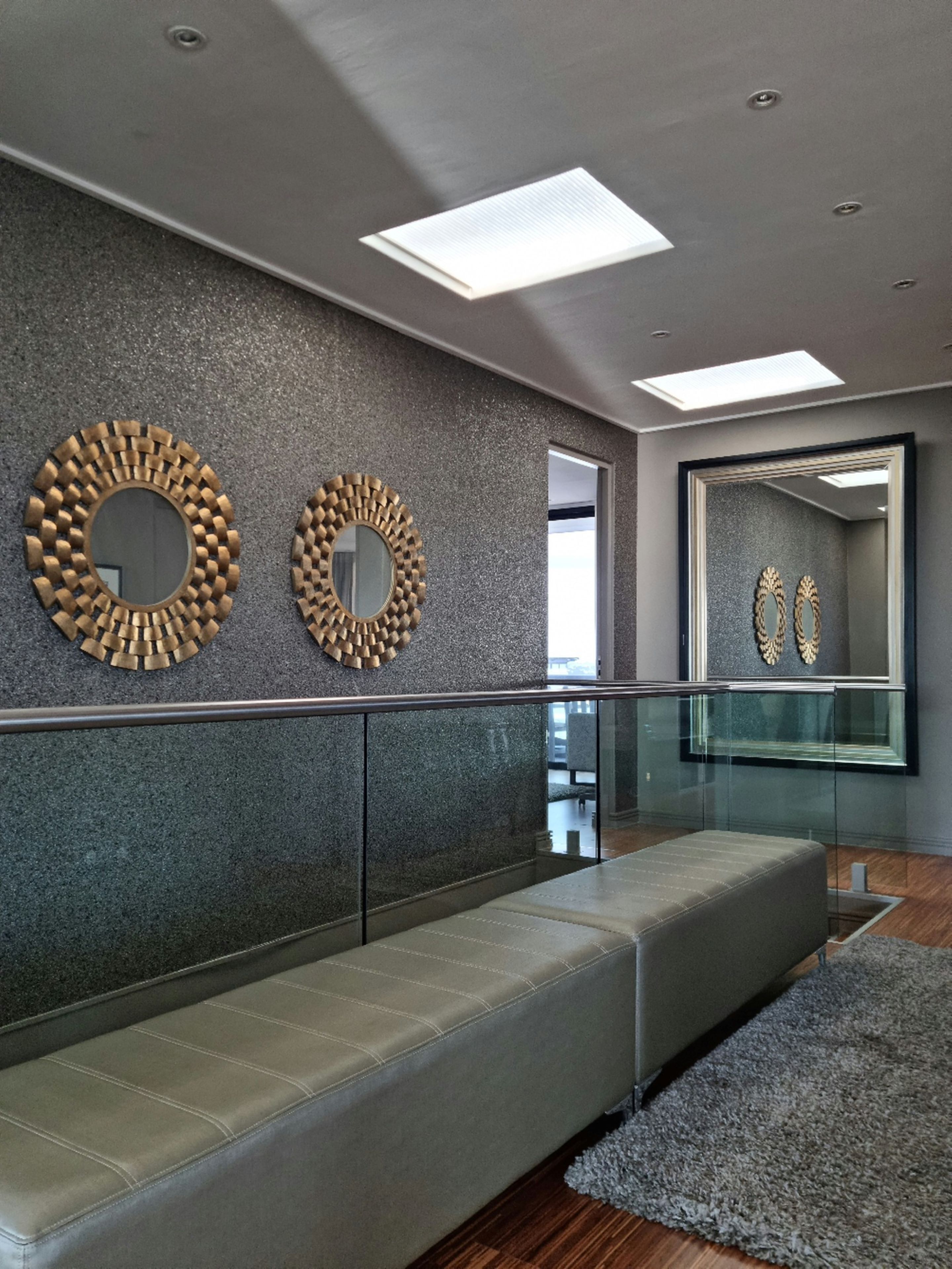 Un intérieur moderne avec un mur en pierre grise, deux miroirs ronds, une rambarde en verre et un banc rembourré de couleur grise.