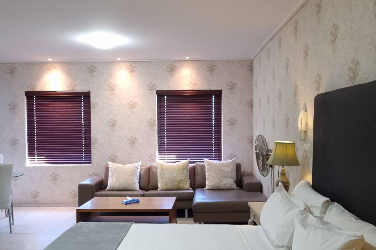 A hotel room with a bed, two sofas, a coffee table, and two windows with blinds.