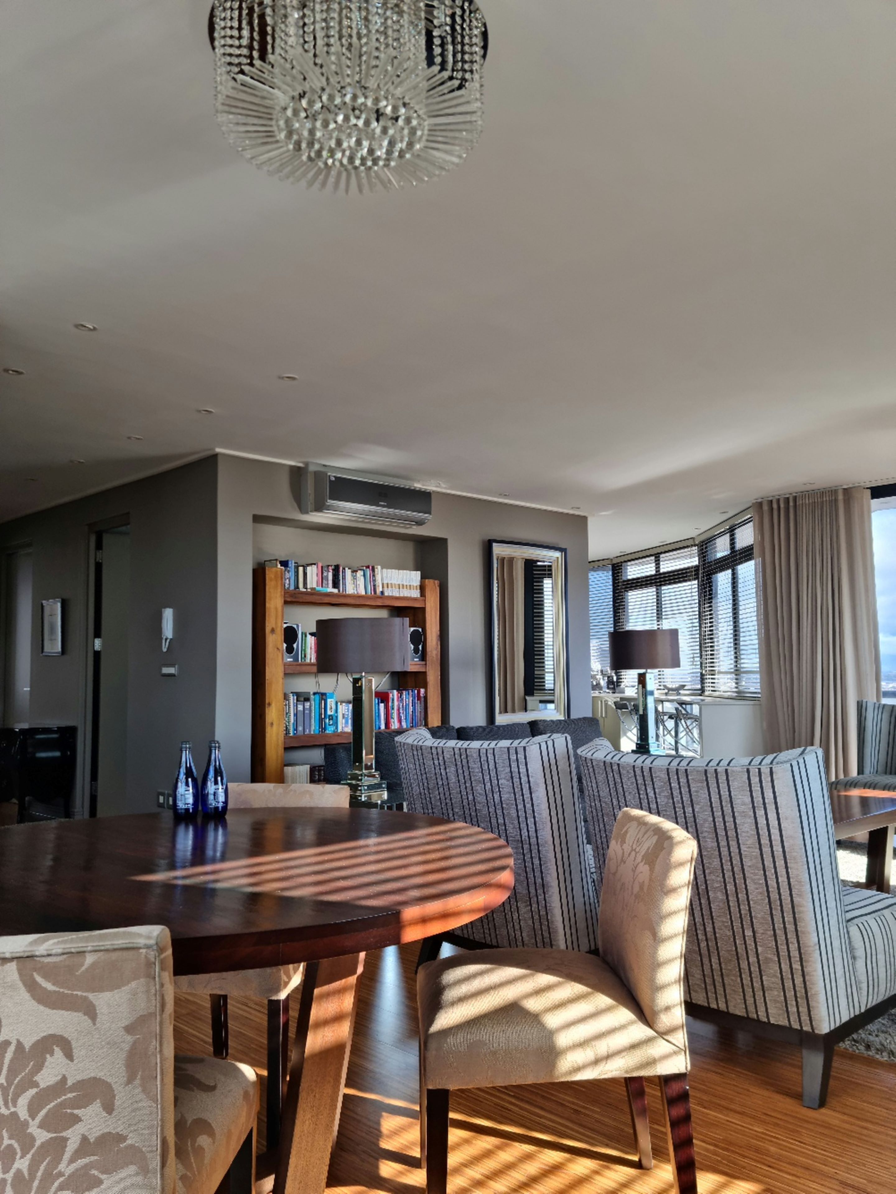 A modern living room with a wooden dining table, striped sofas, a bookshelf, and a large window with curtains.