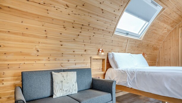 A loft bedroom with a wooden sloped ceiling, a grey sofa with a cushion, a bed with white bedding, and a skylight.