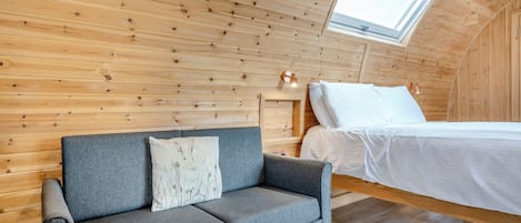 A loft bedroom with a wooden sloped ceiling, a grey sofa with a cushion, a bed with white bedding, and a skylight.