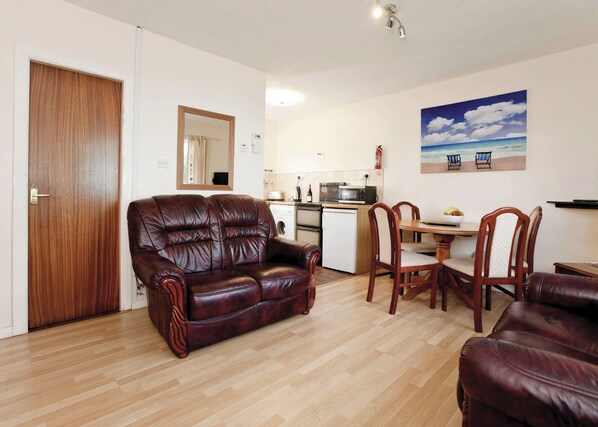 A living room with a leather sofa, wooden dining table, and chairs, a kitchen area with appliances, and a painting of a beach scene. - Bungalow with one double and one single. (Trearddur Bay, Holyhead)