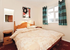 A bedroom with a wooden bed, a bedside table, a nightstand, a mirror, a window with curtains, and a wall-mounted artwork. - Bungalow with one double and one single. (Trearddur Bay, Holyhead)