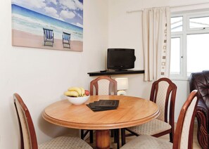 A round wooden dining table with a bowl of fruit, surrounded by chairs, a television on a stand, and a beach painting on the wall. - Bungalow with one double and one single. (Trearddur Bay, Holyhead)