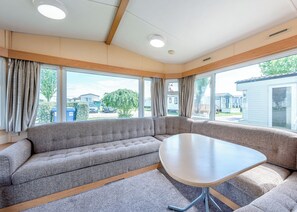 Ein Zimmer mit einer großen Eckcouch, einem runden Tisch und großen Fenstern mit Blick auf eine Wohngegend.
