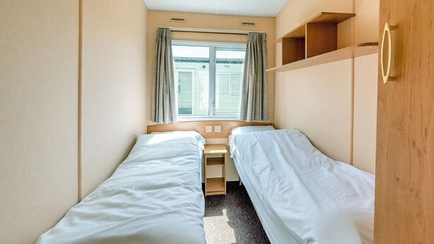 Une petite chambre d’hôtel composée d’une seule pièce, avec deux lits simples, une fenêtre avec des rideaux, une étagère en bois et un luminaire au plafond.