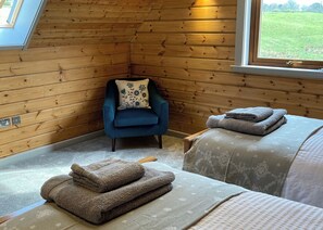 A corner room with wooden walls, a blue chair with a floral pillow, a bed with folded towels, and a window with a view of a green field. - Stunning lodge built on the edge of the lake. (Anglesey)