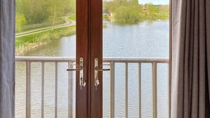Vista a través de una puerta de madera con manija metálica, que da a un balcón con vistas a un río tranquilo, vegetación y cielo despejado.
