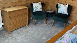 Una habitación acogedora con paredes de madera, un aparador de madera, dos sillones tapizados en verde con cojines de motivos de helechos y una pequeña mesa redonda.