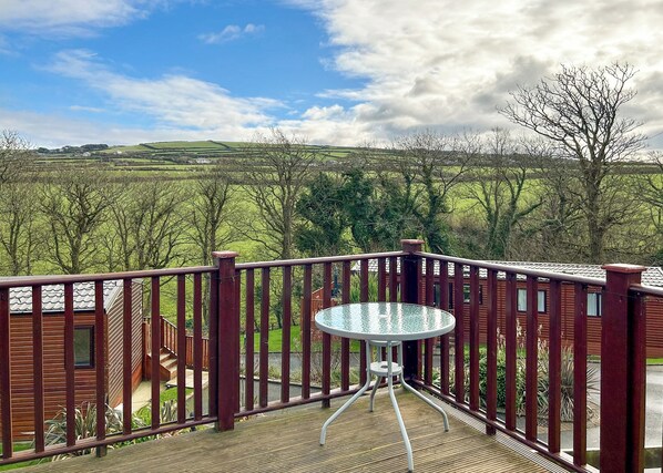 A deck with a glass table, surrounded by a wooden railing, overlooking a green landscape with hills and trees. - Country Holiday Home Four (Tintagel)