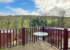 Una terraza con una mesa de vidrio, rodeada por barandas de madera, con vistas a un paisaje verde con colinas y árboles.